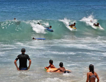 a group of people watches four surfers ride a wave on their stomachs