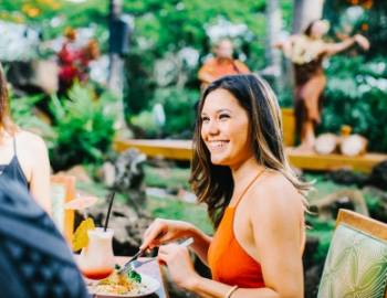 a woman in an orange dress smiles as she eats at keoki's paradise in poipu